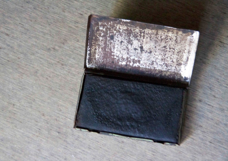 Peut inclure: Une petite bo&icirc;te rectangulaire en m&eacute;tal avec un couvercle &agrave; charni&egrave;re. La bo&icirc;te est ouverte, r&eacute;v&eacute;lant un bloc rectangulaire noir &agrave; l'int&eacute;rieur. La bo&icirc;te a une finition argent&eacute;e us&eacute;e.