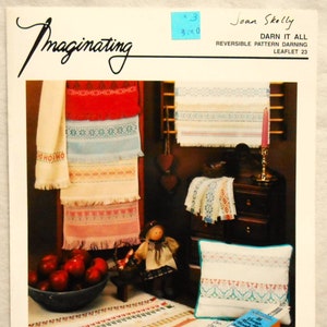 May include: A vintage craft pattern booklet titled "Darn It All" by Joan Skelly. The image showcases various embroidered textiles, including towels and a pillow, with colorful geometric and floral designs. A bowl of red apples and a doll are also visible.