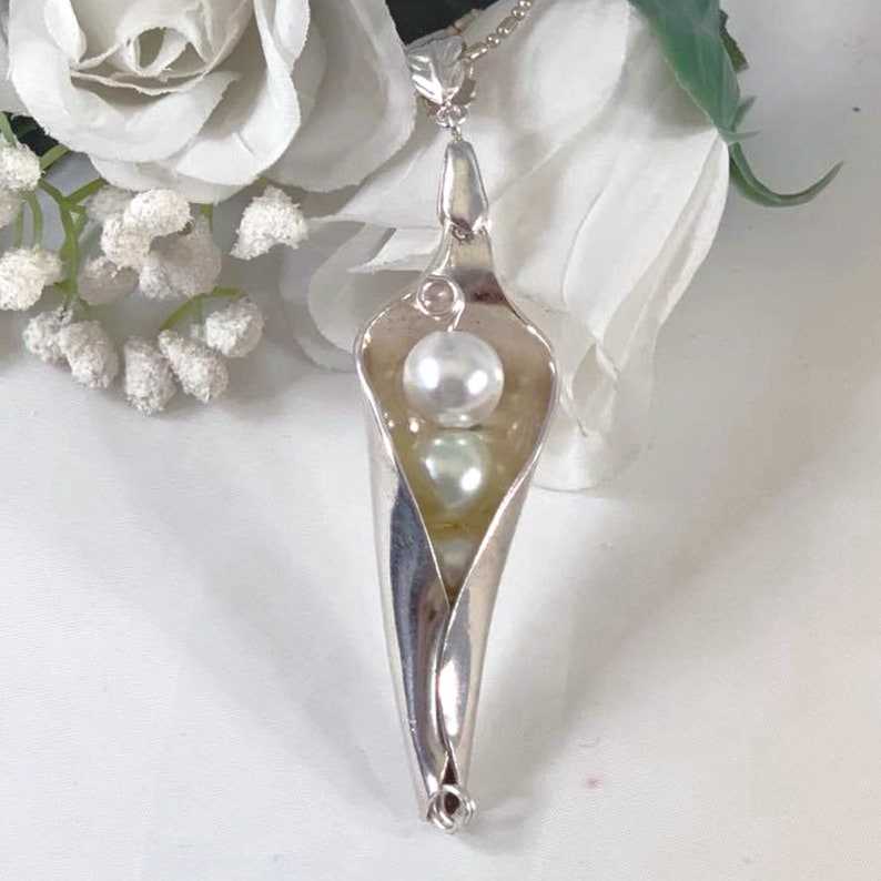 May include: A silver pendant necklace featuring a pea pod design with two white pearls inside. The pendant is suspended from a silver chain.