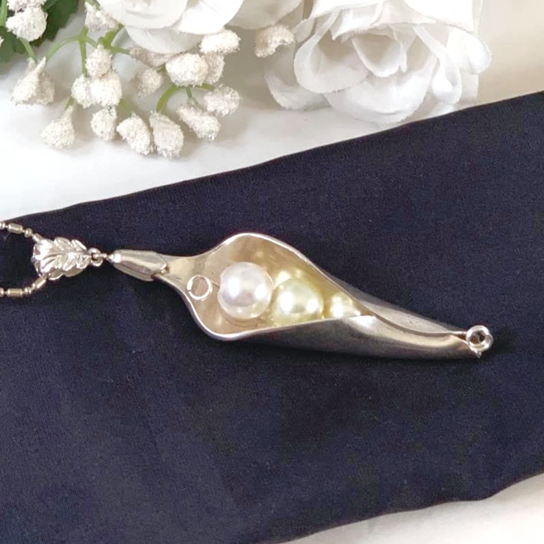 May include: A silver necklace with a pea pod charm. The charm is filled with white and cream colored pearls.