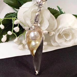 May include: A silver pea pod pendant necklace with three white pearls inside. The necklace is displayed on a white and black surface with white flowers.