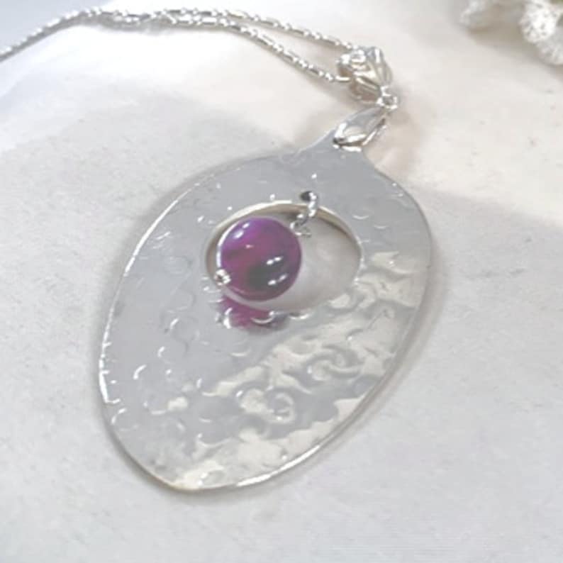 Puede incluir: Un collar de plata con un colgante ovalado martillado con un c&iacute;rculo recortado. Una piedra de &aacute;gata p&uacute;rpura cuelga del colgante.