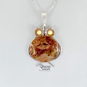 May include: A silver chain necklace with a copper owl pendant. The owl has a round body with a textured surface and yellow eyes. The pendant is attached to the chain with a silver bail. The necklace is labeled "Twisted Silver by Design."