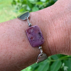 May include: A silver bracelet with a purple and white agate stone charm. The bracelet has a unique design with silver links and a silver clasp.