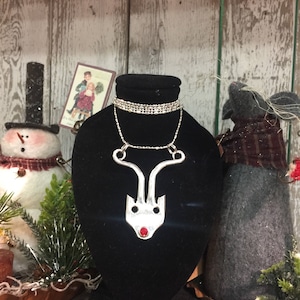 May include: A silver reindeer necklace with a red jewel for a nose. The necklace is displayed on a black velvet stand.