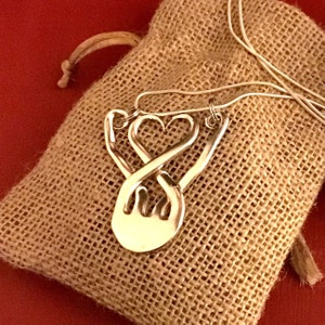 May include: A silver necklace with a pendant shaped like two hands intertwined, forming a heart. The necklace is in a burlap pouch.