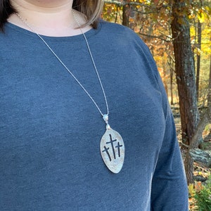 Puede incluir: Un collar de plata con un colgante con forma de cuchara con tres cruces recortadas. El collar lo lleva una persona que lleva una camisa gris. La persona est&aacute; de pie en una zona boscosa.