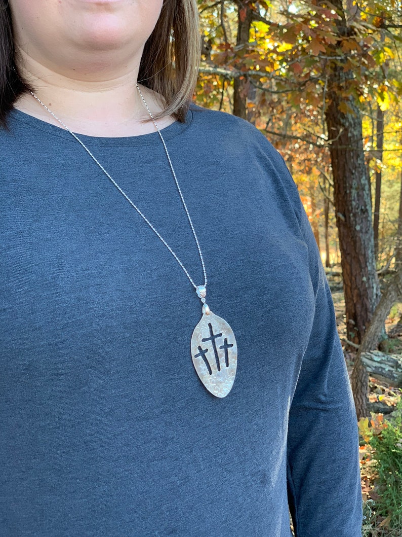 Puede incluir: Un collar de plata con un colgante con forma de cuchara con tres cruces recortadas. El collar lo lleva una persona que lleva una camisa gris. La persona est&aacute; de pie en una zona boscosa.