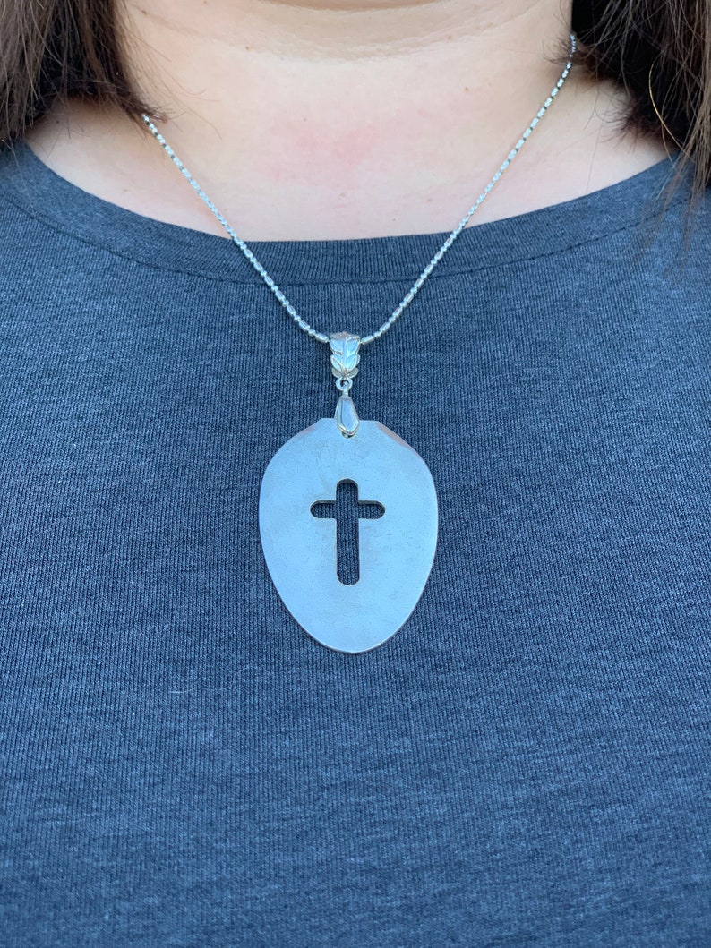 Puede incluir: Un collar de cadena de plata con un colgante con forma de cuchara con una cruz recortada en el centro.
