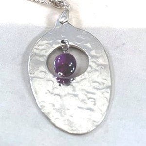 Puede incluir: Un collar de plata con un colgante ovalado martillado que presenta una piedra preciosa redonda de color p&uacute;rpura en un c&iacute;rculo recortado.