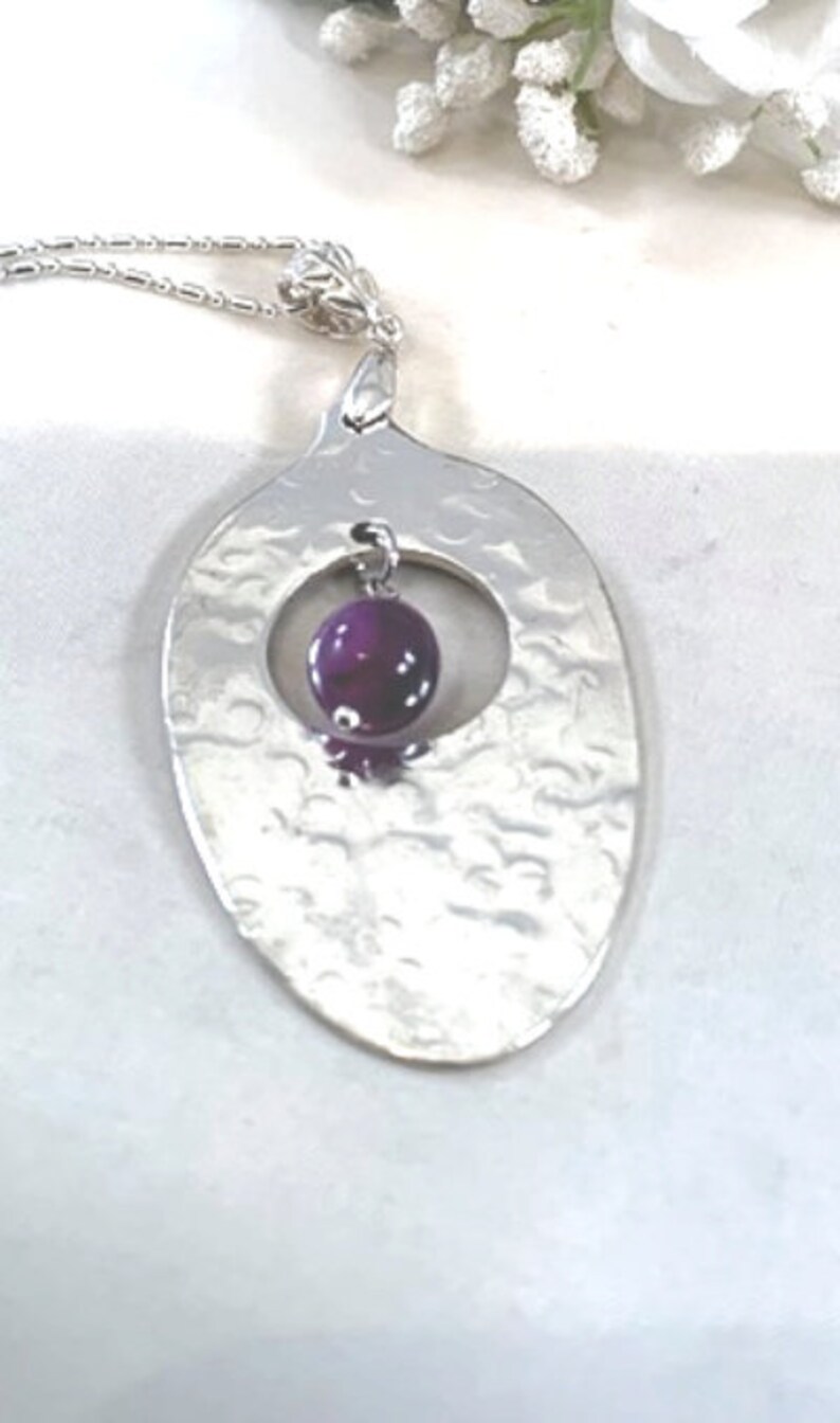 Puede incluir: Un collar de plata con un colgante ovalado martillado que presenta una piedra preciosa redonda de color p&uacute;rpura en un c&iacute;rculo recortado.