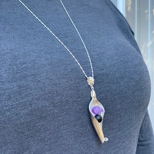 May include: A silver chain necklace with a pendant shaped like a pea pod. The pod is open and contains two purple and one black gemstone.
