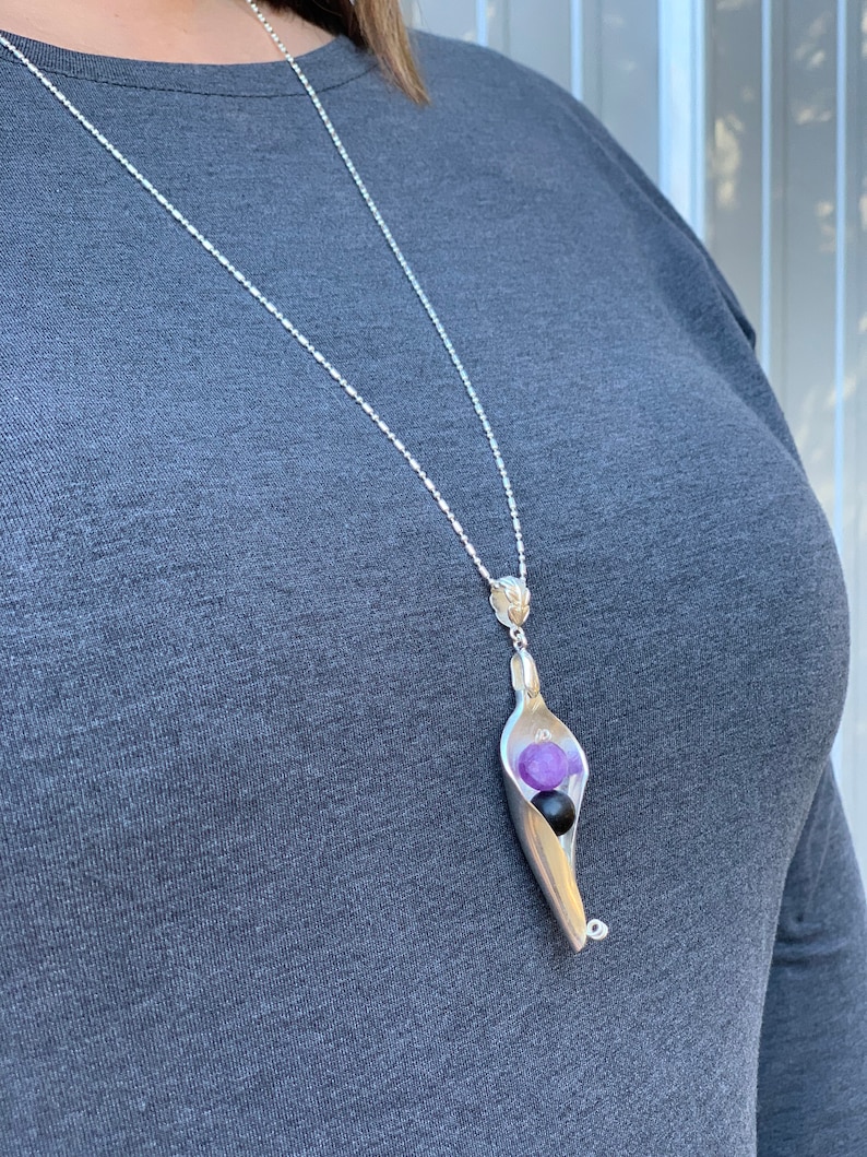 May include: A silver chain necklace with a pendant shaped like a pea pod. The pod is open and contains two purple and one black gemstone.