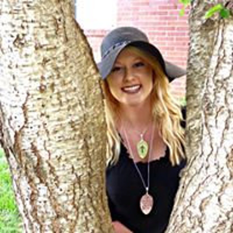 Puede incluir: Una mujer con un sombrero negro y una blusa negra con dos collares de plata. Los collares tienen un colgante en forma de hoja con un dise&ntilde;o de cruz.
