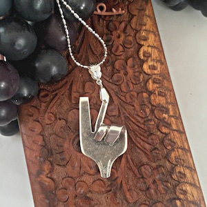 May include: A silver necklace with a peace sign pendant made from a bent fork. The pendant is on a wooden surface with a carved design.