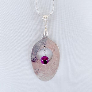 Puede incluir: Un collar con un colgante de cuchara de plata y una cuenta de &aacute;gata p&uacute;rpura que cuelga del centro. La cuchara ha sido martillada para crear una superficie texturizada y tiene una abertura circular en el centro.