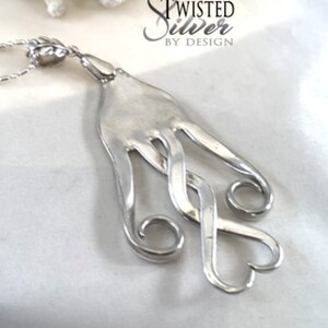 May include: A silver necklace with a pendant made from a twisted fork. The pendant is shaped like two hearts. The necklace is on a white background. Twisted Silver by Design.