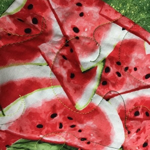 May include: A close-up of a red and green fabric with a watermelon print. The fabric is quilted and has a white background with black seeds.