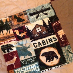 May include: A colorful patchwork quilt with a variety of nature-themed designs, including bears, moose, cabins, and fishing. The quilt features the text "Great Outdoors", "One with Nature", "Wild Times", "Candle and Kayak", "Welcome to the Cabin", "Fishing", "Cabin", "Moo", "Get Back", "Bear Country", and "Task Wild".