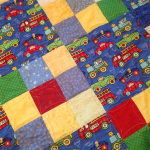 May include: A blue, yellow, green, and red patchwork quilt with a pattern of toy cars and trucks. The quilt is made of squares and rectangles.
