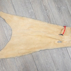 May include: A light beige fur pelt with a small hole in the center. The fur is soft and fluffy, and the pelt is laid out flat on a wooden surface.