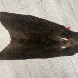 May include: A dark brown fur pelt with a small red tag attached to it. The fur is soft and fluffy, and the pelt is laid out flat on a wooden surface.