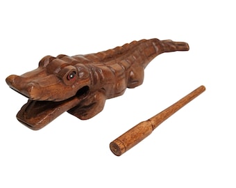 May include: Wooden alligator hand percussion instrument with a brown wooden stick. The alligator has a carved mouth that opens and closes when the stick is used to strike the instrument.