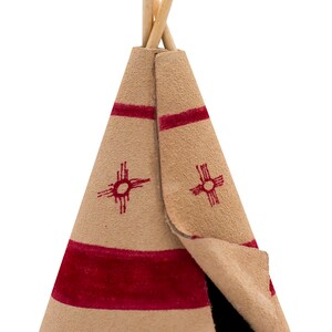 May include: A small, tan leather teepee with red trim and a red sun symbol. The teepee has two wooden poles sticking out of the top.