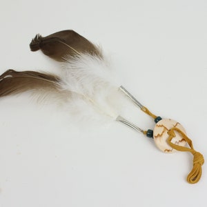May include: A decorative hair accessory featuring two brown and white feathers attached to a light-colored, oval-shaped pendant. The pendant is adorned with a leather cord and small beads, creating a rustic, bohemian aesthetic.