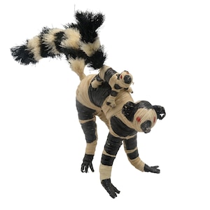 May include: A black and white lemur with a long, bushy tail, made of woven straw. The lemur is carrying a smaller lemur on its back.