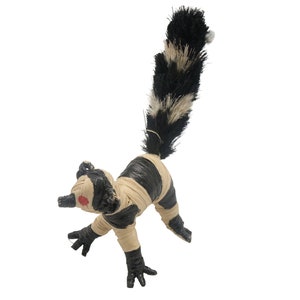 May include: A black and white striped stuffed animal with a long, bushy tail. The animal has a red dot on its face and is made of straw.