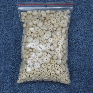 May include: A plastic bag filled with a variety of white wooden beads. The beads are of different shapes and sizes, including round, oval, and cylindrical.