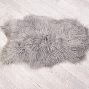 May include: A soft, gray sheepskin rug with long, fluffy fur. The rug is laid out flat on a light wood floor.