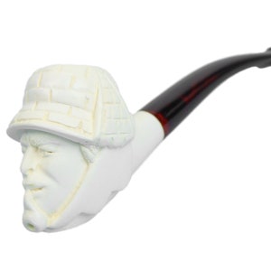 May include: White ceramic pipe with a sculpted human face wearing a hat. The pipe has a brown stem.