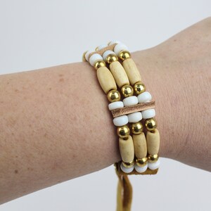 May include: A multi-strand bracelet with wooden beads, white beads, and gold beads. The bracelet has a brown leather cord.