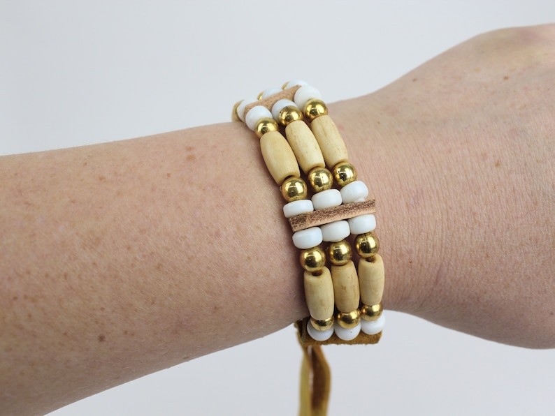 May include: A multi-strand bracelet with wooden beads, white beads, and gold beads. The bracelet has a brown leather cord.