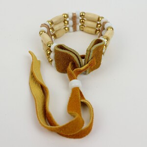 May include: A triple-strand beaded bracelet with wooden beads, gold beads, and white beads. The bracelet has a brown leather tie closure.