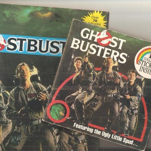May include: Two vintage Ghostbusters items are displayed. One is a book with the Ghostbusters logo and images of the cast. The other item is a box with the Ghostbusters logo and the text "12 Collector Stickers Inside".