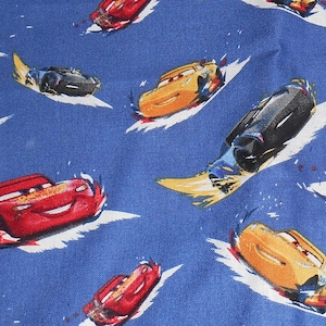 May include: Blue fabric featuring a pattern of cartoon race cars in red, yellow, and black. The cars are shown with speed lines, indicating movement. Ideal for children's clothing or nursery decor.