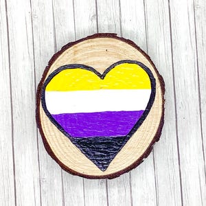May include: A wooden round slice with a painted heart in the colors of the non-binary pride flag. The heart is yellow, white, purple, and black.