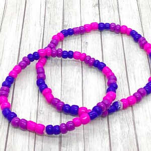 May include: Two beaded bracelets in shades of pink, purple, and blue. The bracelets are made of small, round beads and are arranged on a light wood-grain surface. The bracelets are a vibrant accessory.