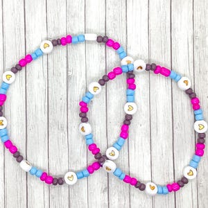 May include: Two friendship bracelets made with colorful beads. The bracelets are made with pink, blue, brown, and white beads. Each bead has a small gold heart on it.