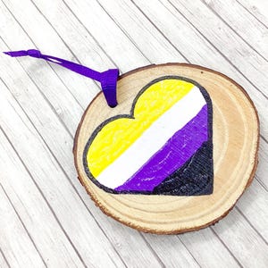 Puede incluir: Adorno de madera con forma de corazón que muestra los colores de la bandera del orgullo no binario: amarillo, blanco, morado y negro. El adorno tiene una cinta morada para colgar y está hecho de una rodaja de madera natural.
