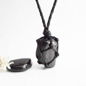 May include: Black gemstone pendant necklace with a black cord. The gemstone is wrapped in a macrame knot design. The necklace is displayed on a white background with a small black stone next to it.
