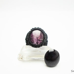 May include: A black ring with a purple gemstone set in a woven design. The ring is displayed on a white crystal and a black stone.