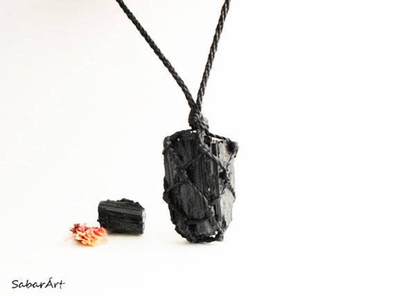 May include: Black tourmaline crystal pendant wrapped in black cord, hanging from a black cord necklace. The pendant is approximately 2 inches long. There is a smaller black tourmaline crystal next to the pendant.