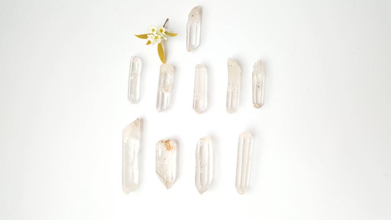 May include: A collection of ten clear quartz crystal points arranged in a symmetrical pattern on a white background. The crystals are various sizes and shapes, with some having a slight golden hue.