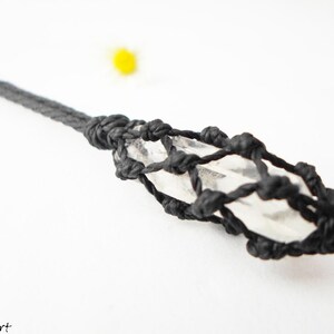 May include: A clear quartz crystal is wrapped in a black cord, creating a knotted, geometric design. The crystal is suspended from a black cord.