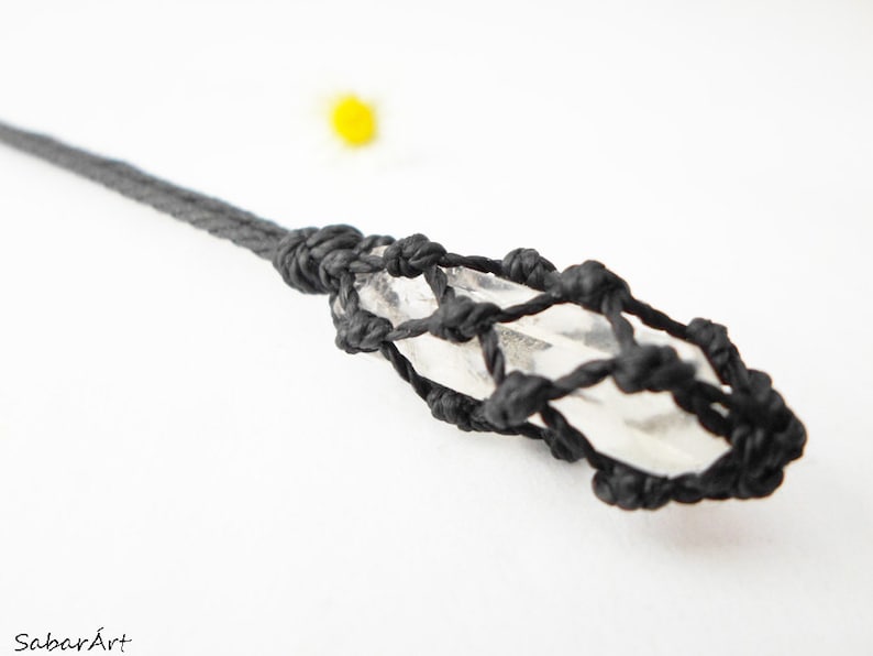 May include: A clear quartz crystal is wrapped in a black cord, creating a knotted, geometric design. The crystal is suspended from a black cord.