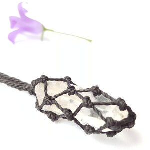 May include: A clear quartz crystal is wrapped in a black macrame cord, creating a pendant necklace. The crystal is long and slender, with a pointed tip. The macrame cord is tied in a intricate pattern, creating a delicate and elegant design.